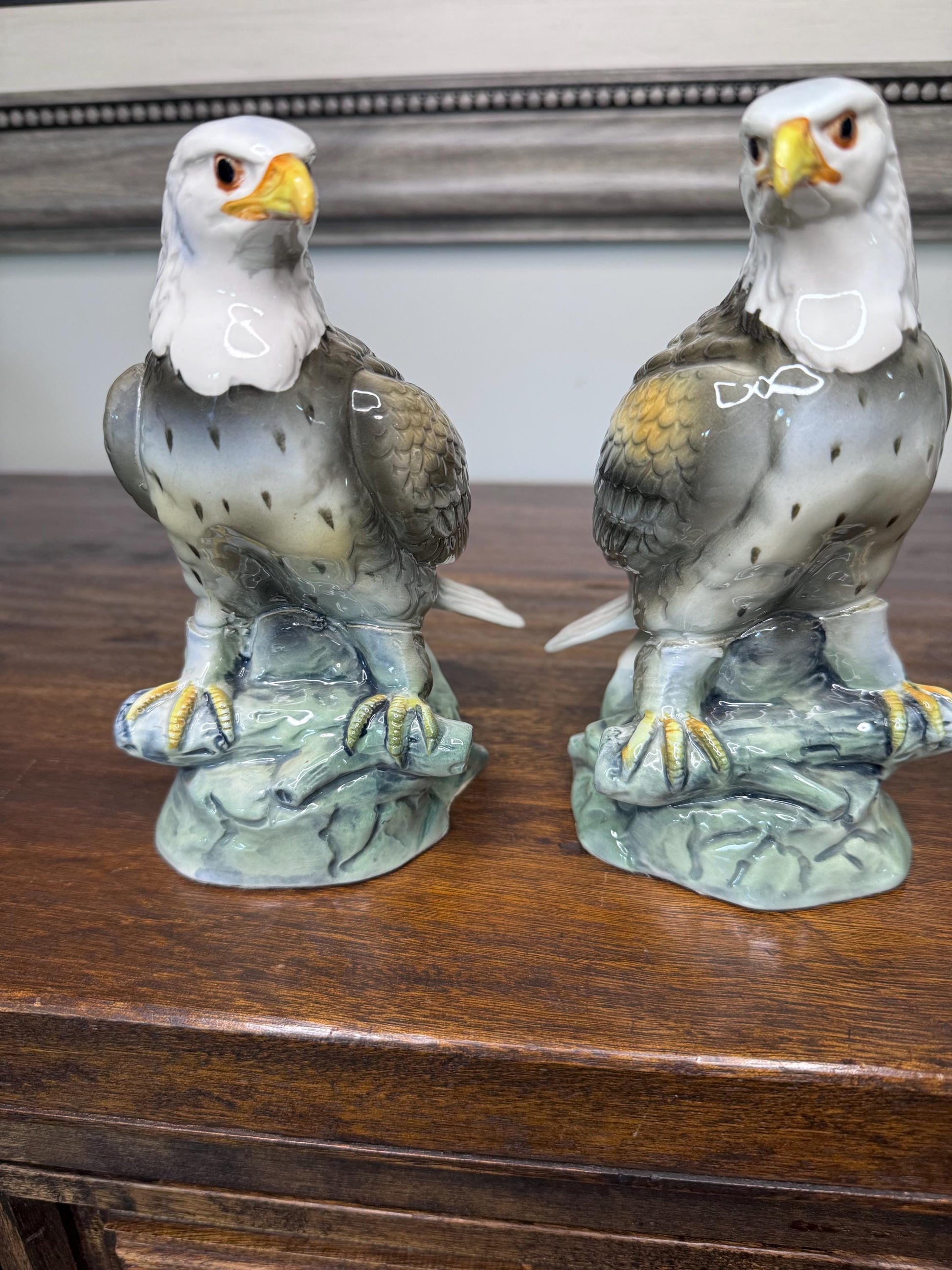 May include: Two porcelain eagle figurines perched on green bases. The eagles have white heads, yellow beaks, and brown and gray bodies with yellow accents. The figurines are on a wooden surface.