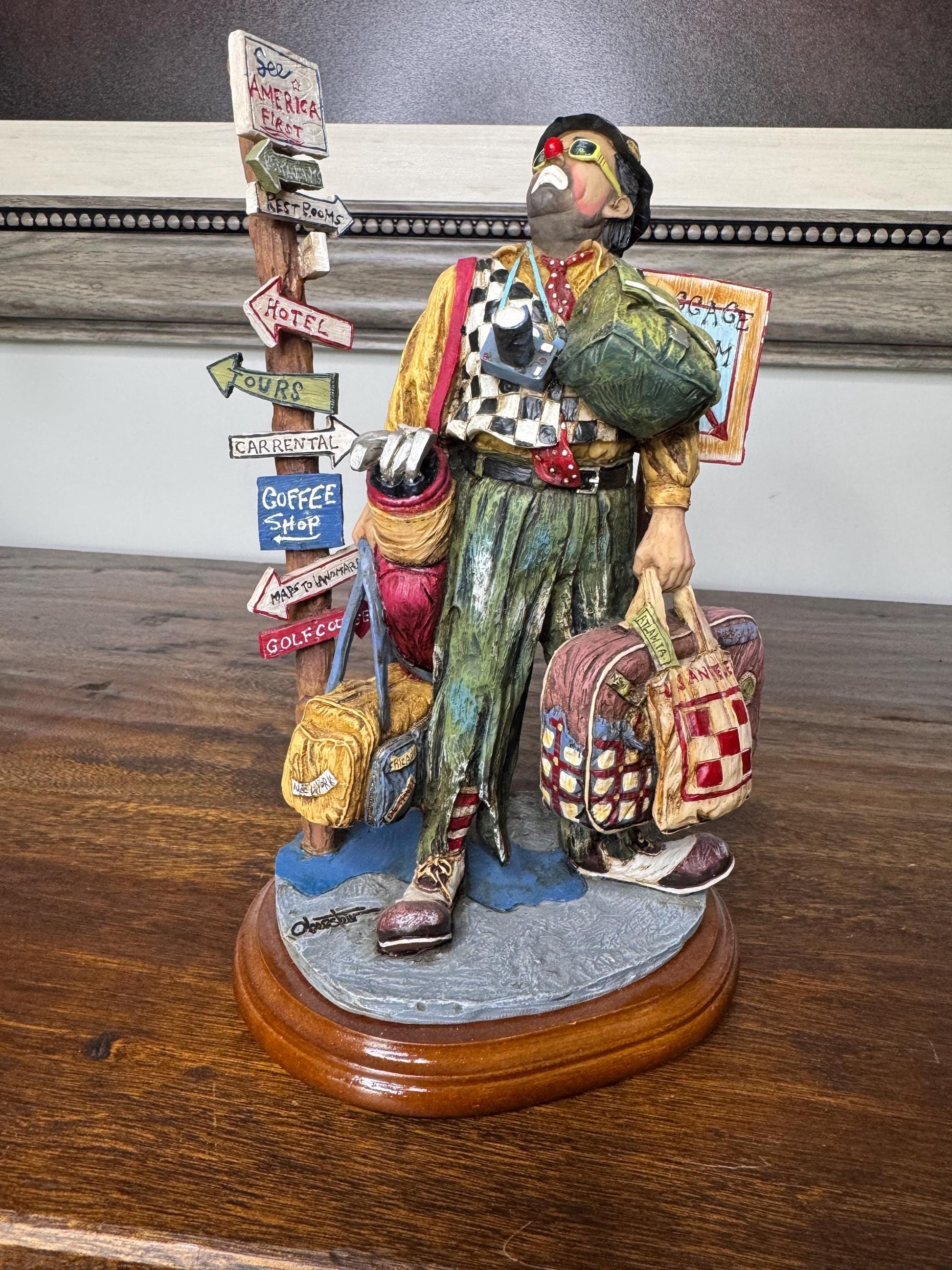 May include: A clown figurine with a red nose and green glasses, wearing a checkered vest and holding a camera and multiple suitcases. The figurine is standing next to a wooden signpost with arrows pointing to various destinations, including &#39;See America First&#39;, &#39;Rest Rooms&#39;, &#39;Hotel&#39;, &#39;Tours&#39;, &#39;Car Rental&#39;, &#39;Coffee Shop&#39;, &#39;Maps to Anywhere&#39;, and &#39;Golf Course&#39;. The figurine is standing on a wooden base.