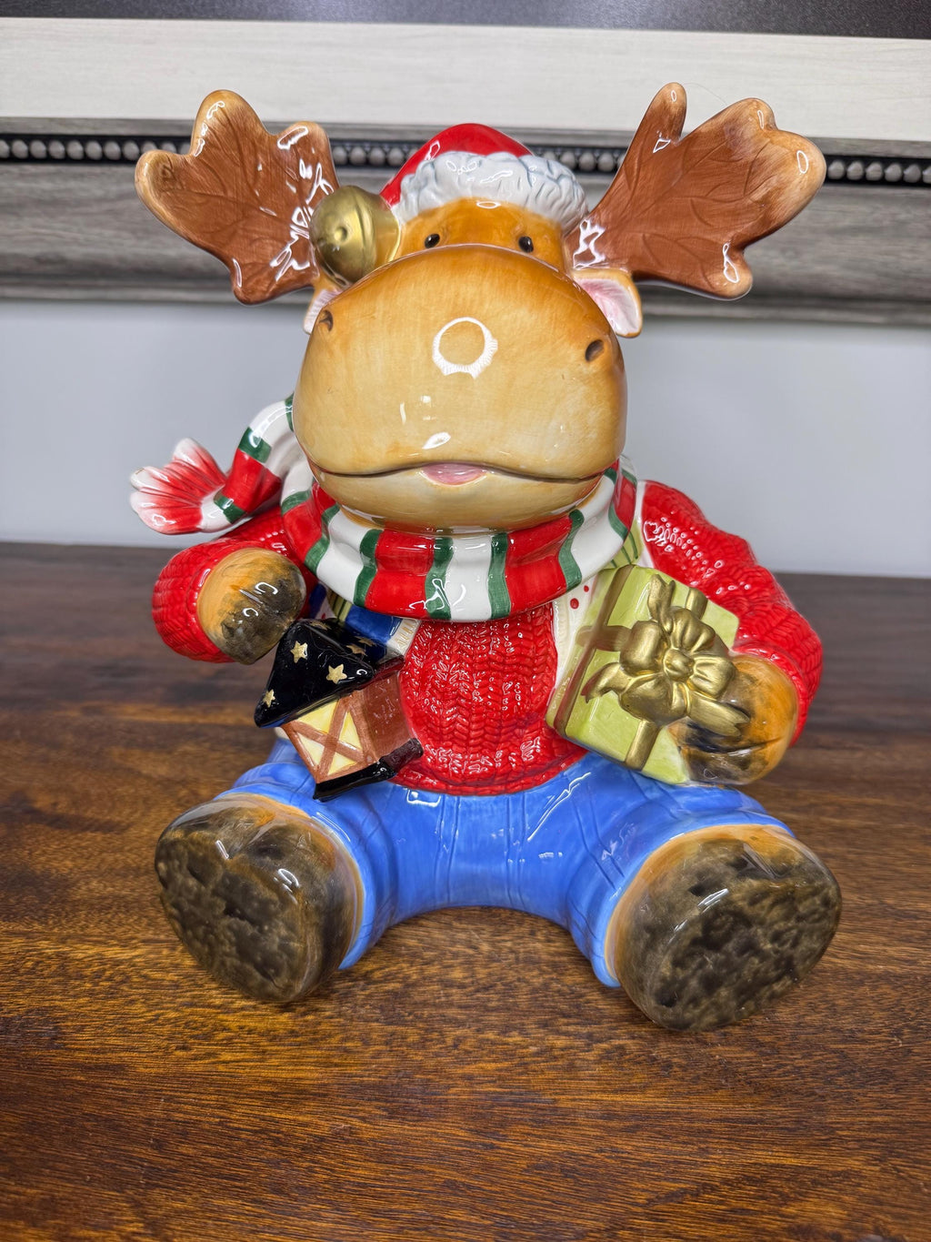May include: A ceramic Christmas moose figurine wearing a red sweater, blue jeans, and a green and red striped scarf. The moose is holding a small house and a wrapped gift. The moose has a Santa hat on its head and bells around its neck.
